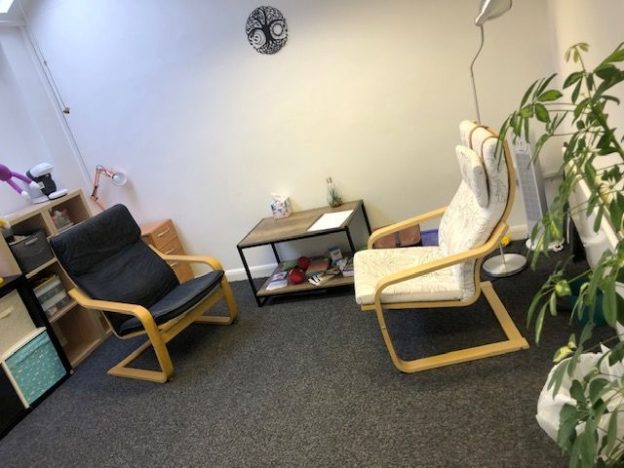 A calm counselling room with two comfortable chairs facing each other, soft lighting, and plants creating a relaxed, welcoming space.